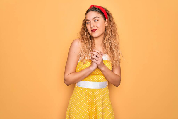 Beautiful blonde pin-up woman with blue eyes wearing diadem standing over yellow background laughing nervous and excited with hands on chin looking to the side - Fotoğraf, Görsel