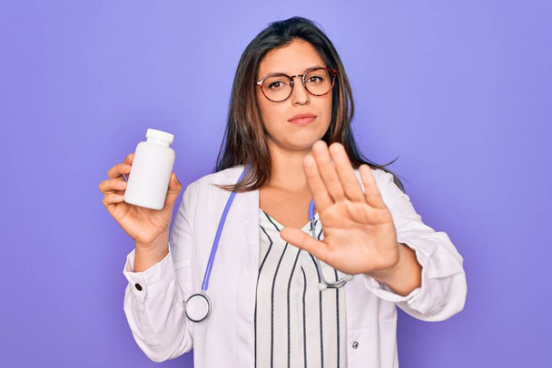 Young professional doctor woman holding pharmaceutical pills over purple background with open hand doing stop sign with serious and confident expression, defense gesture - Photo, Image