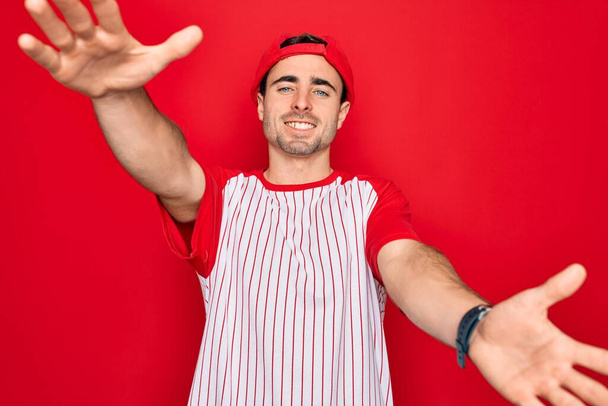 Young handsome sporty man with blue eyes wearing striped baseball t-shirt and cap looking at the camera smiling with open arms for hug. Cheerful expression embracing happiness. - Photo, Image