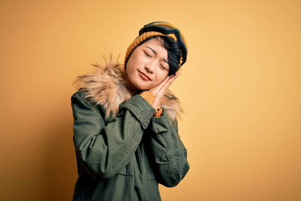 Gyönyörű ázsiai síelő lány visel hó sportruházat segítségével síszemüveg sárga háttér alszik fáradt álmodozás és pózol kéz együtt, miközben mosolyog csukott szemmel. - Fotó, kép