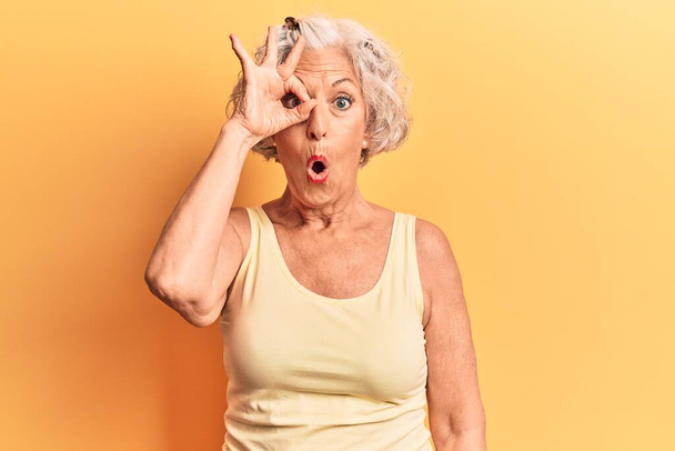 Starsza siwowłosa kobieta w luźnych ciuchach robi ok gest wstrząśnięty zaskoczoną twarzą, okiem patrzącym przez palce. niewierząca ekspresja.  - Zdjęcie, obraz