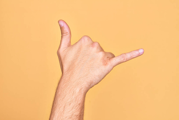 Ruka kavkazského mladíka ukazující prsty na izolovaném žlutém pozadí gestikulující havajským gestem na pozdrav, telefonem a komunikačním symbolem - Fotografie, Obrázek