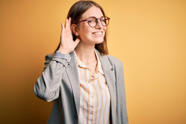 Young beautiful redhead woman wearing jacket and glasses over isolated yellow background smiling with hand over ear listening an hearing to rumor or gossip. Deafness concept. - Fotografie, Obrázek