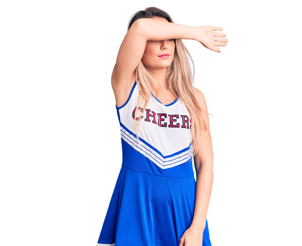 Young beautiful blonde woman wearing cheerleader uniform covering eyes with arm, looking serious and sad. sightless, hiding and rejection concept  - Foto, Imagem