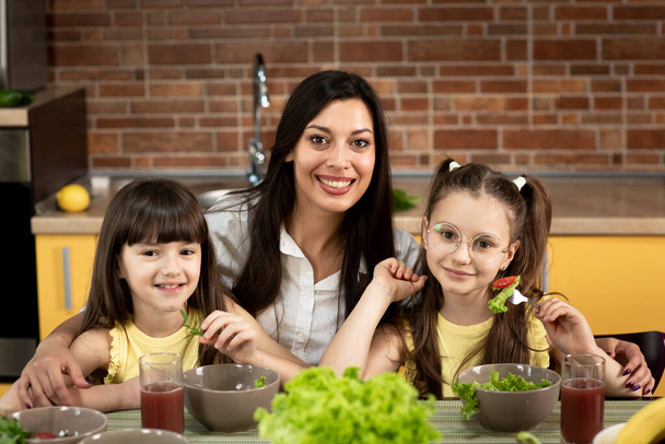 Mãe alegre e duas filhas estão comendo salada saudável juntos em casa. O conceito de alimentação saudável, valores familiares, tempo juntos
 - Foto, Imagem