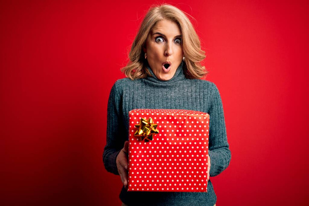 Средневековая красивая блондинка с подарком на день рождения на изолированном красном фоне, напуганная от удивления лицом, напуганная и взволнованная выражением страха
 - Фото, изображение