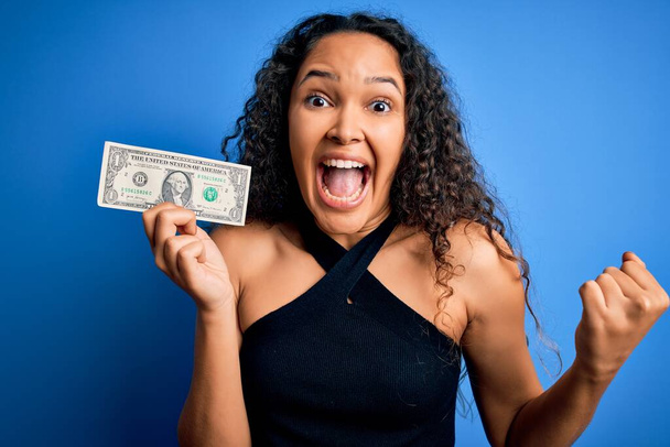 Mladá krásná žena s kudrnaté vlasy drží jeden dolar nad izolované modré pozadí křičí hrdý a slaví vítězství a úspěch velmi vzrušený, povzbuzující emoce - Fotografie, Obrázek