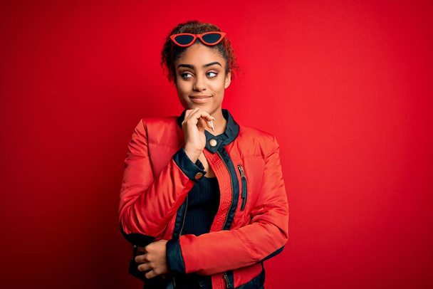 Beautiful african american girl wearing red jacket and sunglasses over isolated background with hand on chin thinking about question, pensive expression. Smiling with thoughtful face. Doubt concept. - Fotoğraf, Görsel
