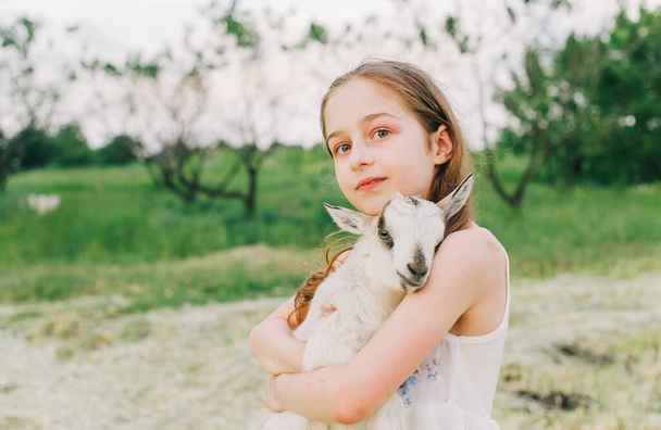 girl with baby goat on farm outdoors. Love and care. Village animals. happy child hugs goat, concept of unity of nature and man. Friendship of child and animals. Happy childhood. Girl and goat - Photo, Image