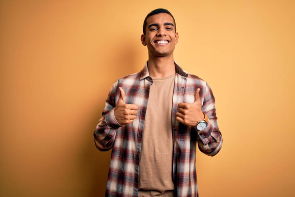 Junger gutaussehender afrikanisch-amerikanischer Mann in lässigem Hemd, das über gelbem Hintergrund steht und eine positive Geste mit der Hand macht, Daumen hoch lächelnd und glücklich. Fröhliche Miene und Siegergeste. - Foto, Bild