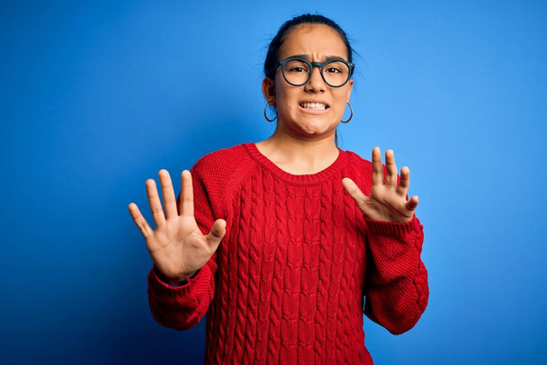 Junge schöne asiatische Frau in lässigem Pullover und Brille über blauem Hintergrund angewidert Ausdruck, unzufrieden und ängstlich dabei Ekel Gesicht, weil Abneigung Reaktion. Mit erhobenen Händen - Foto, Bild