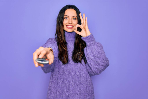Jeune belle femme utilisant la télécommande de télévision sur fond violet isolé faisant signe ok avec les doigts, excellent symbole - Photo, image