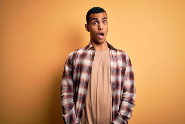 Young handsome african american man wearing casual shirt standing over yellow background In shock face, looking skeptical and sarcastic, surprised with open mouth - 写真・画像
