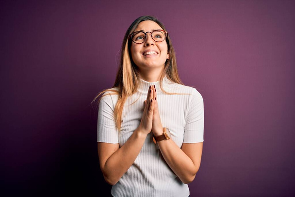 Young beautiful blonde woman with blue eyes wearing casual t-shirt over purple background begging and praying with hands together with hope expression on face very emotional and worried. Begging. - Photo, Image