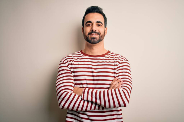 Young handsome man with beard wearing casual striped t-shirt standing over white background happy face smiling with crossed arms looking at the camera. Positive person. - Фото, зображення
