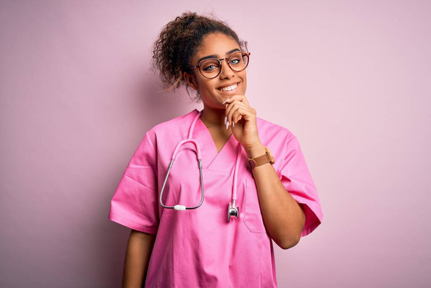 医療用の制服を着たアフリカ系アメリカ人看護婦の女の子とピンクの背景の上に聴診器を身に着けているアフリカ系アメリカ人看護師は、交差した腕と手をあごに上げ笑顔でカメラを見て自信を持っている。肯定的な考え. - 写真・画像