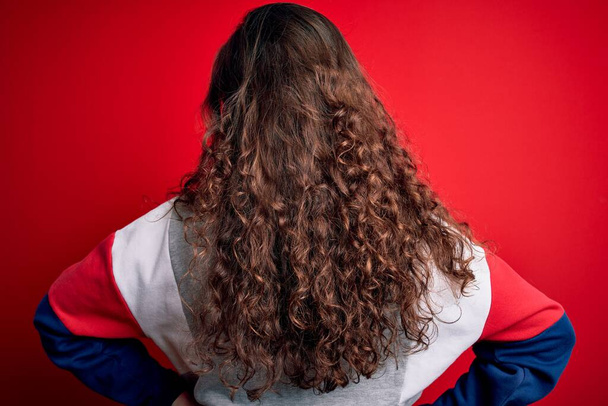 Nuori kaunis nainen kihara tukka yllään rento huppari yli eristetty punainen tausta seisoo taaksepäin katsellen pois kädet kehon - Valokuva, kuva