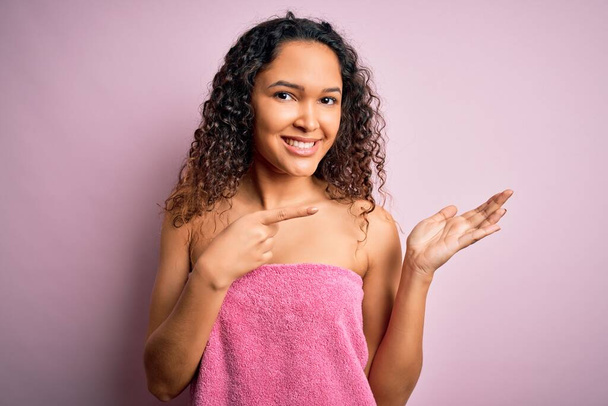 Mladá krásná žena s kudrnaté vlasy nosí sprchový ručník po koupeli přes růžové pozadí ohromen a usmívá se na kameru při prezentaci s rukou a ukazuje prstem. - Fotografie, Obrázek