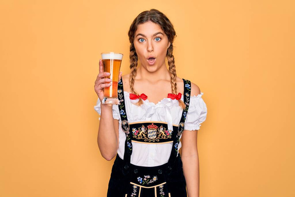 Красивая блондинка немецкая женщина с голубыми глазами в платье Октоберфест пьет стакан пива испуганный от шока с удивленным лицом, напуганный и взволнованный выражением страха
 - Фото, изображение