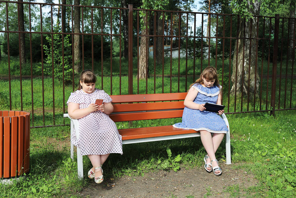 Chicas jóvenes regordetas están sentadas en un banco del parque a distancia social. Están ocupados con sus dispositivos y no se comunican entre sí. El problema del exceso de peso en los niños después de la cuarentena
 - Foto, Imagen