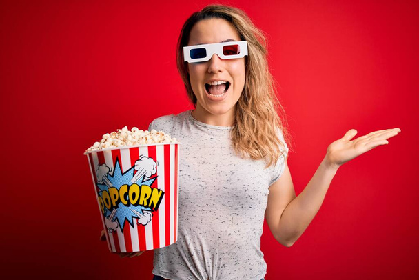 Nuori kaunis blondi nainen katsomassa elokuvaa käyttäen 3D-lasit syöminen popcorn välipala erittäin onnellinen ja innoissaan, voittaja ilme juhlii voittoa huutaen iso hymy ja nosti kädet - Valokuva, kuva