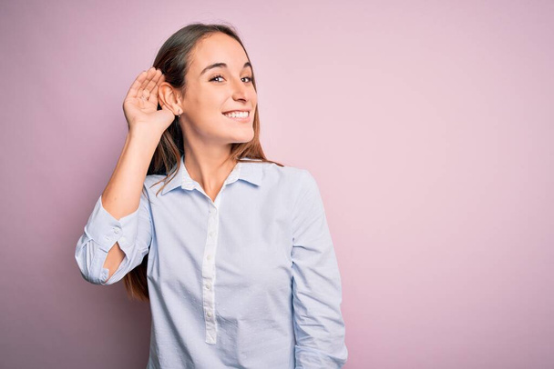 Young beautiful businesswoman wearing elegant shirt standing over isolated pink background smiling with hand over ear listening an hearing to rumor or gossip. Deafness concept. - Фото, изображение