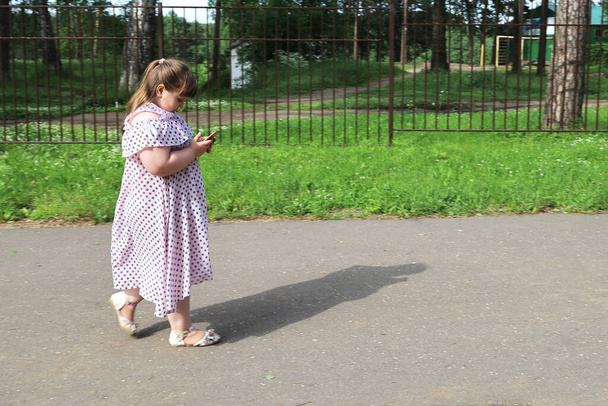 Kleines, dickes Mädchen läuft einen verlassenen Parkweg entlang, schaut auf ein Smartphone und denkt nicht an Gefahren. Das Konzept der Sucht von Kindern nach elektronischen Geräten - Foto, Bild