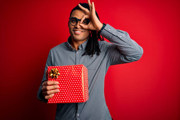 Афроамериканец с дредами держит коробку на красном изолированном фоне со счастливым лицом улыбается делает хорошо знак с рукой на глазу глядя сквозь пальцы
 - Фото, изображение