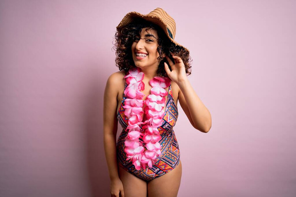 Young beautiful arab woman on vacation wearing swimsuit and hawaiian lei flowers smiling with hand over ear listening an hearing to rumor or gossip. Deafness concept. - Foto, imagen
