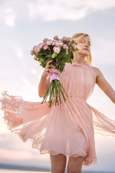 A girl in a delicate pink dress is spinning with a bouquet of pink peony roses. Selective focus. - Foto, afbeelding