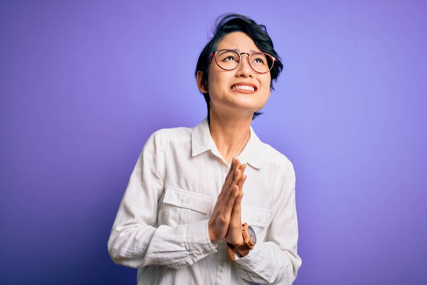 Jong mooi aziatisch meisje dragen casual shirt en bril staan over paarse achtergrond bedelen en bidden met de handen samen met hoop uitdrukking op het gezicht zeer emotioneel en bezorgd. Smeken. - Foto, afbeelding