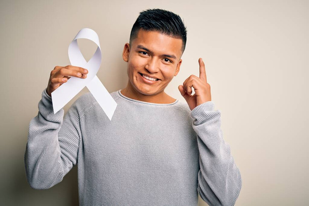 Mladý latinský muž držící bílou stužku proti rakovině zastaví ženy násilí překvapené nápadem nebo otázkou ukazující prstem se šťastnou tváří, číslo jedna - Fotografie, Obrázek