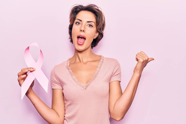 Young beautiful woman holding pink cancer ribbon pointing thumb up to the side smiling happy with open mouth  - Photo, image