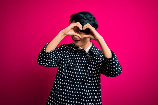 Joven hermosa chica asiática con chaqueta casual de pie sobre fondo rosa aislado haciendo forma de corazón con la mano y los dedos sonriendo mirando a través de signo
 - Foto, imagen