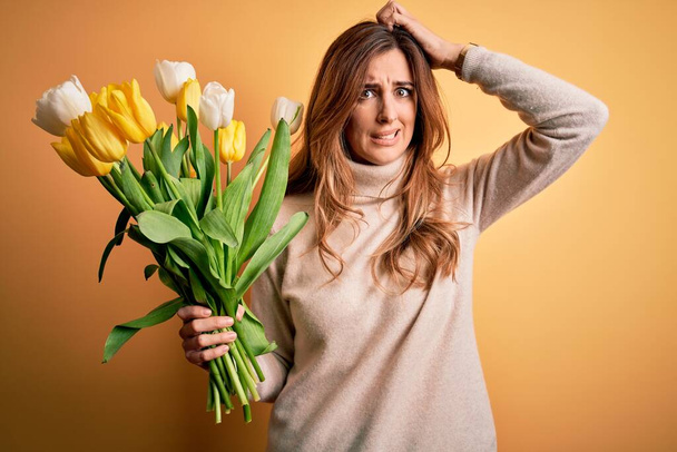Young beautiful brunette woman holding bouquet of yellow tulips over isolated background confuse and wondering about question. Uncertain with doubt, thinking with hand on head. Pensive concept. - Zdjęcie, obraz