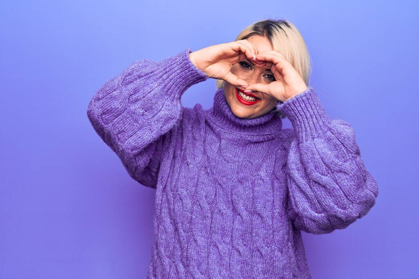 Beautiful blonde plus size woman wearing casual turtleneck sweater over purple background Doing heart shape with hand and fingers smiling looking through sign - Foto, Imagen