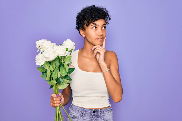 Gyönyörű afro-amerikai nő gazdaság csokor fehér virágok felett lila háttér komoly arc gondolkodás kérdés, nagyon zavaros ötlet - Fotó, kép