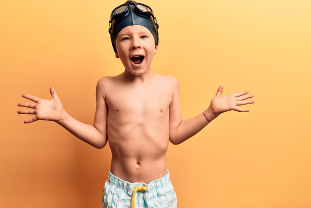 Nettes blondes Kind in Badebekleidung und Schwimmerbrille feiert Sieg mit glücklichem Lächeln und Siegerausdruck mit erhobenen Händen  - Foto, Bild