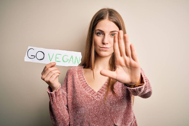 Junge schöne rothaarige Frau, die nach veganem Essen fragt, hält Papier mit veganer Botschaft mit offener Hand und macht Stoppschild mit ernstem und selbstbewusstem Gesichtsausdruck, Verteidigungsgeste - Foto, Bild
