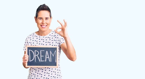 Young woman with short hair holding blackboard with dream word doing ok sign with fingers, smiling friendly gesturing excellent symbol  - Photo, Image