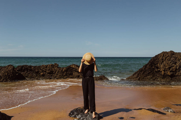 Girl on a background of the sea in black clothes.A girl on a background of the Spanish landscape.A woman on a background of blue ocean.Girl covers her face with a hat on the beach - Fotografie, Obrázek
