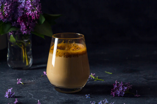 Summer still life with dalgona coffee in tall glass and lilac flowers on dark background. Instant coffee whipped with sugar and water and added to cold milk. Cool summer drink. Copy space for text - Fotografie, Obrázek