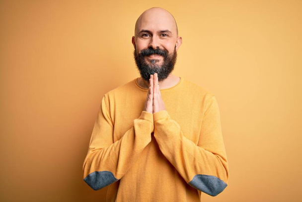 手で一緒に祈って黄色の背景に立ってカジュアルなセーターを身に着けている髭のハンサムなはげ男は自信を持って笑顔許しを求めて. - 写真・画像
