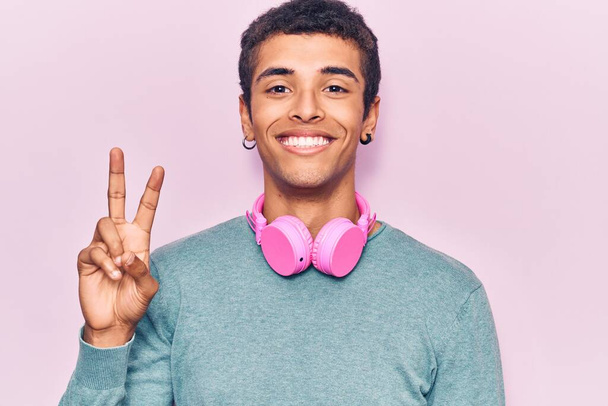 Junger afrikanisch-amerikanischer Mann, der mit Kopfhörern Musik hört, lächelt mit glücklichem Gesicht in die Kamera und macht ein Siegeszeichen. Nummer zwei.  - Foto, Bild