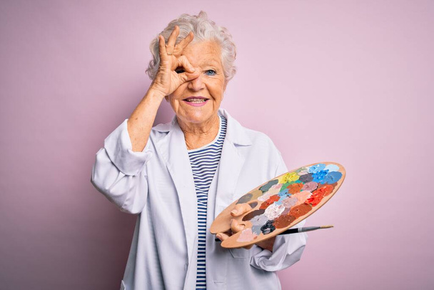 Senior belle artiste aux cheveux gris femme peinture à l'aide d'un pinceau et palette sur fond rose avec le visage heureux souriant faire ok signe avec la main sur l'œil regardant à travers les doigts - Photo, image