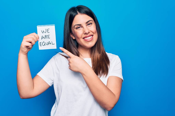 Young beautiful brunette woman asking for equality holding paper with we are equal message smiling happy pointing with hand and finger - Foto, immagini