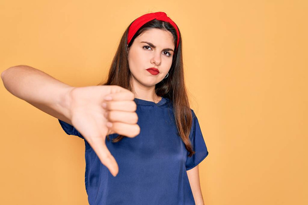 Young beautiful brunette woman wearing red lips over yellow background looking unhappy and angry showing rejection and negative with thumbs down gesture. Bad expression. - Photo, Image