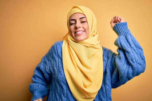 Young beautiful brunette muslim woman wearing arab hijab over isolated yellow background stretching back, tired and relaxed, sleepy and yawning for early morning - Photo, Image