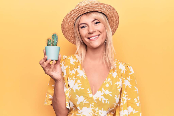 Piękna blondynka nosi letni kapelusz trzymając doniczkę kaktusa na żółtym tle patrząc pozytywnie i szczęśliwy stojąc i uśmiechając się z pewnym uśmiechem pokazując zęby - Zdjęcie, obraz