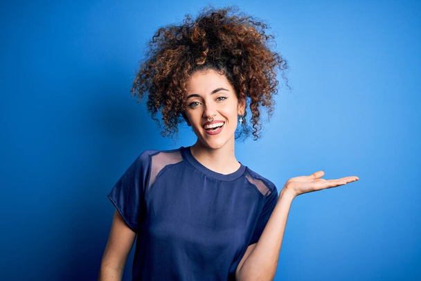 Mladá krásná žena s kudrnatými vlasy a piercing na sobě ležérní modré tričko s úsměvem veselý prezentaci a ukazuje dlaní ruky při pohledu do kamery. - Fotografie, Obrázek